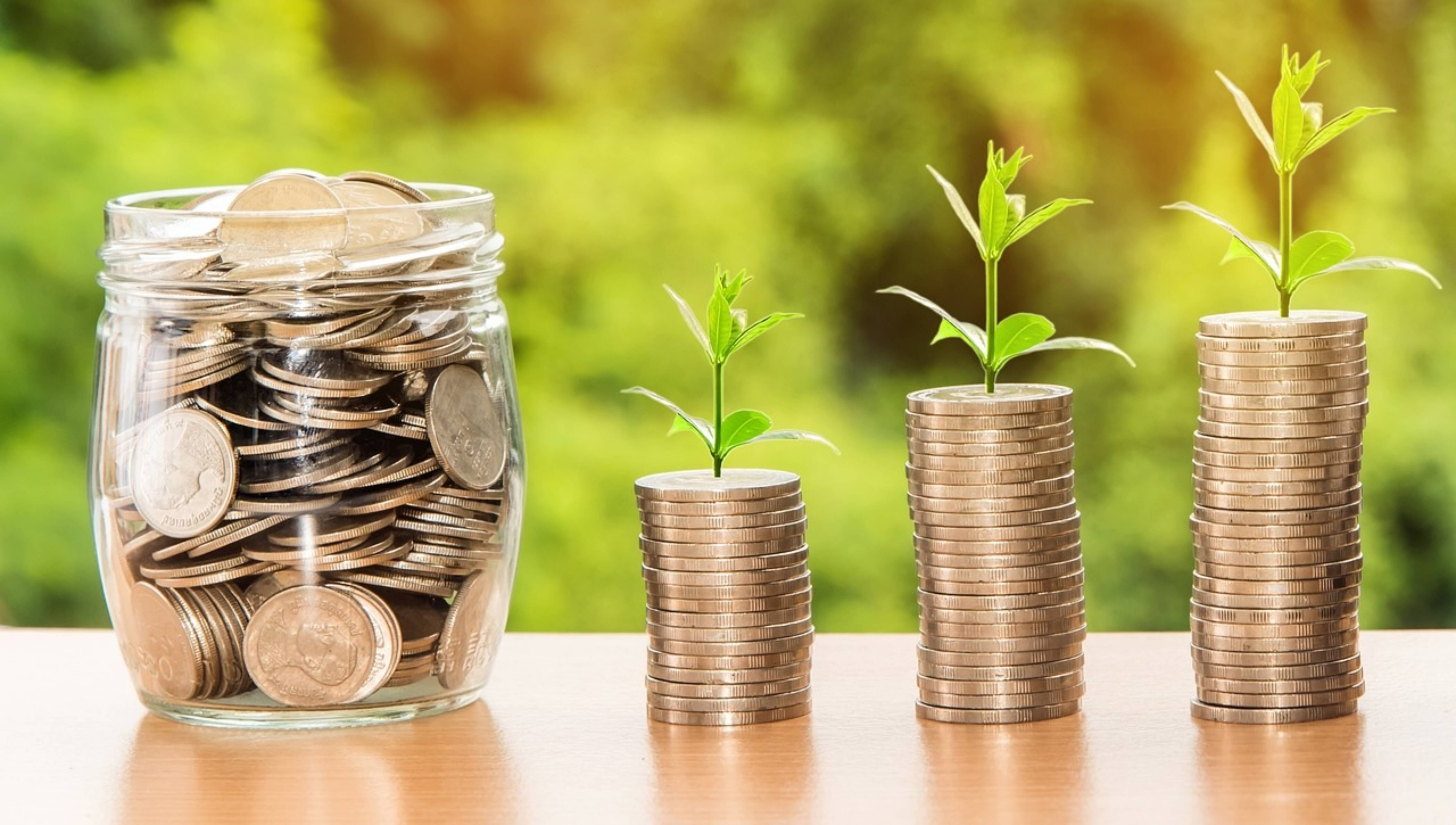 A glass jar filled with coins next to stacks of coins sprouting from the top.