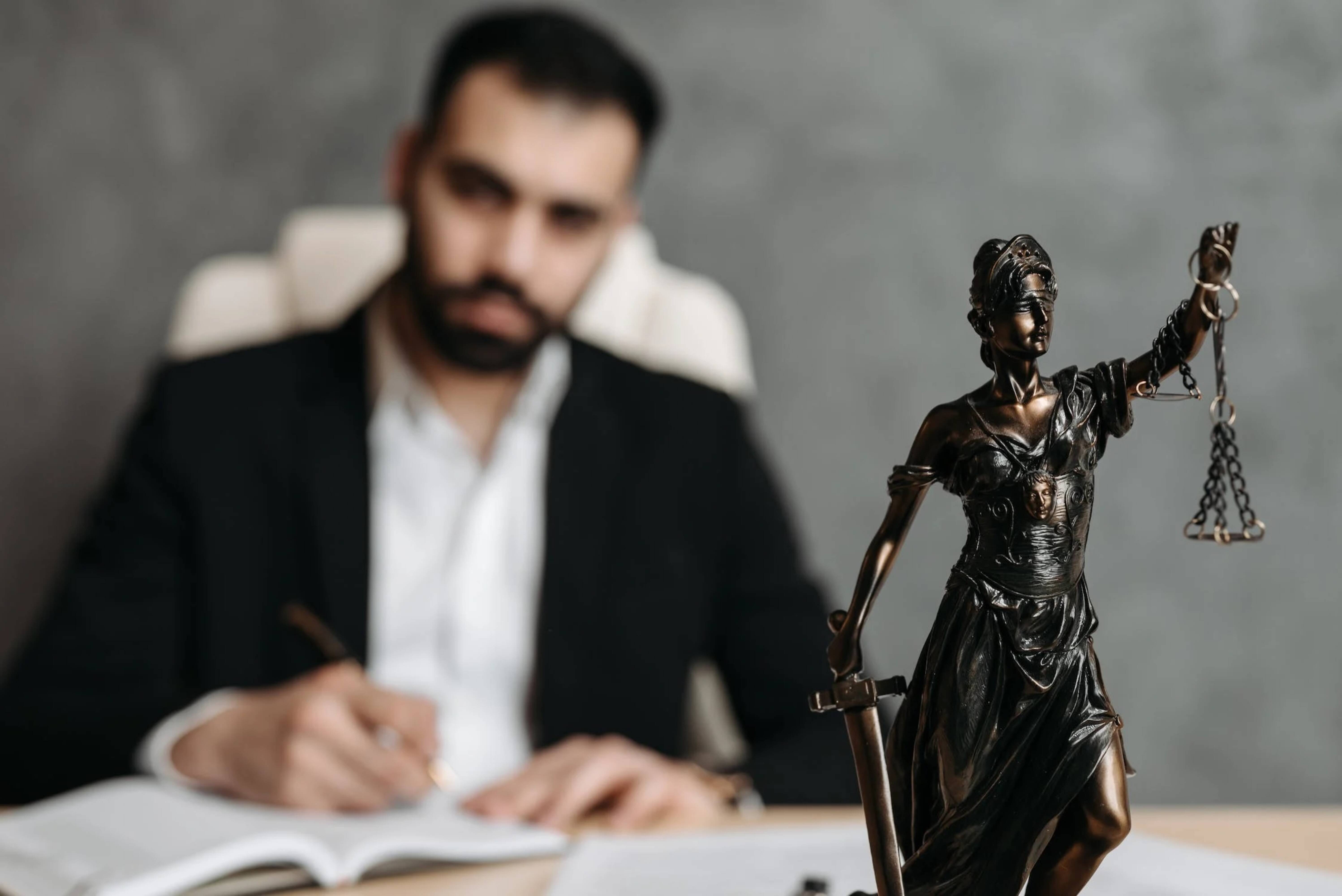 A man in a suit signing a contract behind a statue of Lady Justice.